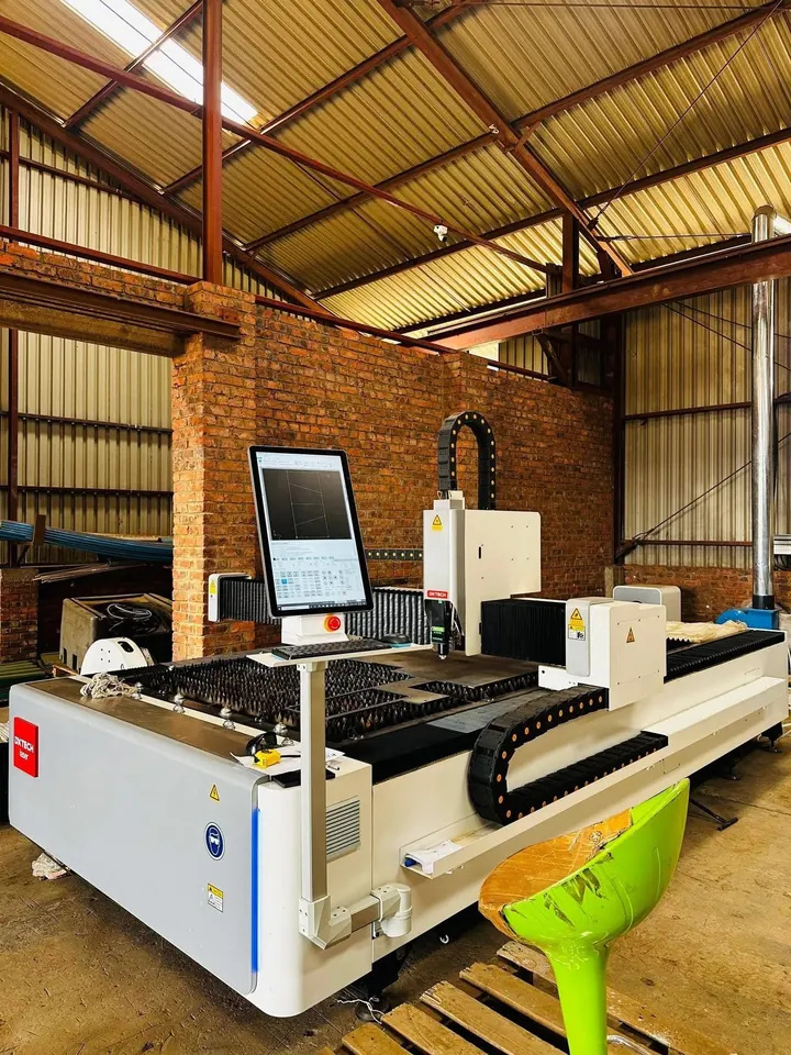A fibre laser cutting machine in a workshop, with a brick pier, yellow sheeting and a control screen beside the bed.