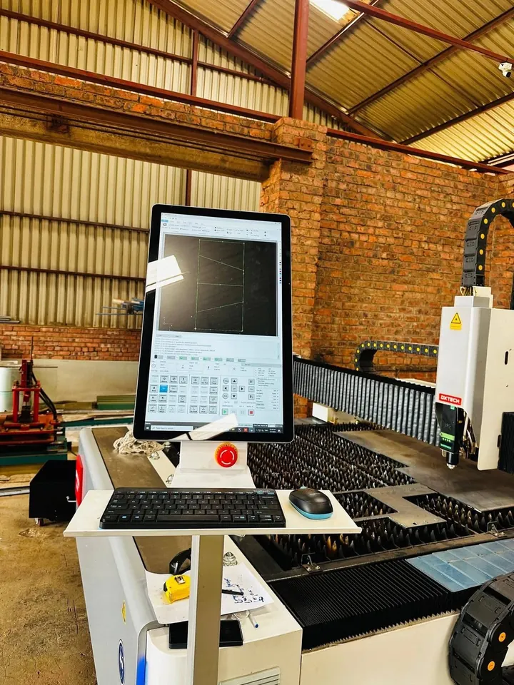The laser control station: an upright monitor showing a nested cutting path on screen, keyboard, mouse and emergency stop on the machine shelf, with the slatted bed and the DXTECH cutting head behind it.