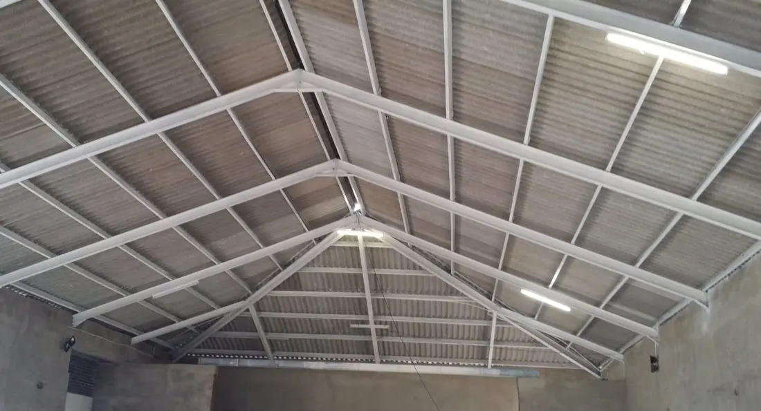 A white-painted roof structure seen from inside: trusses, purlins and corrugated sheeting with rooflights between the bays.
