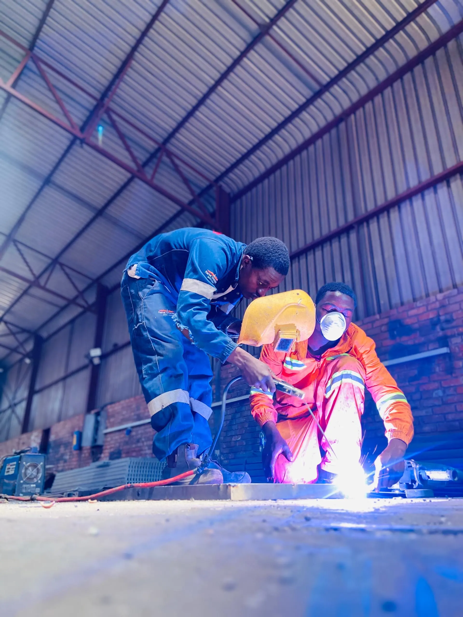 Two Kingson Engineering workers welding a steel grating panel on the floor of the Harare workshop, seen from low down with the workshop roof trusses and sheeting above them.